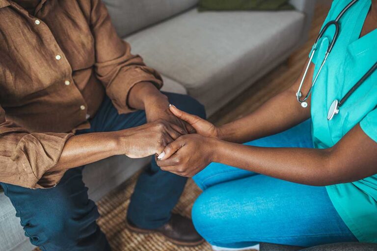 An adult child sits beside their elderly parent in a cozy assisted living room, engaging in a gentle conversation.